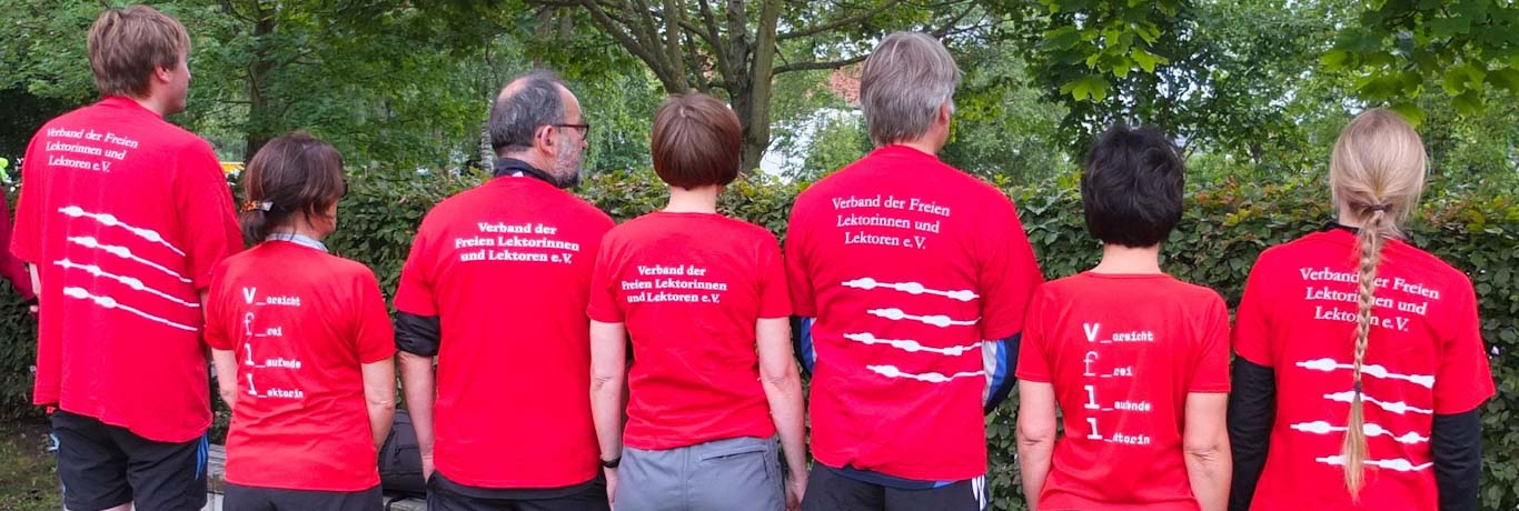 7 Menschen stehen nebeneinander, mit dem Rücken zur Kamera. So sieht man die unterschiedlichen Texte auf ihren roten T-Shirts. Alle haben das VFLL-Logo, aber in unterschiedlicher Ausführung. Bei manchen steht die Abkürzung VFLL für "Vorsicht frei laufende LektorIn