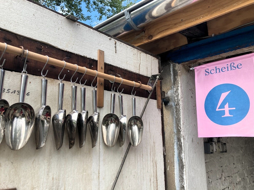 An einem Stab hängen lauter kleine silberne Handschaufeln, daneben ein rosa Schild mit einer großen blauen 4 darauf und der Überschrift "Scheiße".