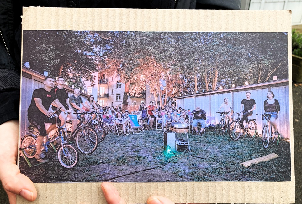 Ein Foto wurde auf Pappkarton geklebt und zeigt offensichtlich einen Filmabend mitten in den Garagen, bei dem die Energie für den gezeigten Film mit umstehenden Fahrrädern erzeugt wird.