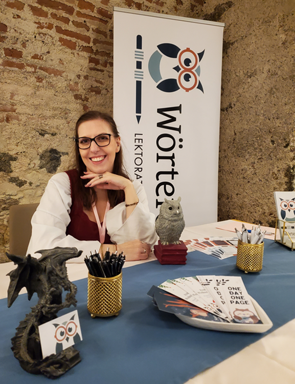 Foto von der Lektorin Rebekka Haindl vor einem Banner mit ihrem Logo der "Wörteule". Sie sitzt an ihrem Stand, der dekoriert ist mit Wörtereulen-Visitenkarten, gehalten von einem Drachen. Auf dem Tisch steht auch die Figur einer Eule, es liegen Lesezeichen und Stifte aus. 