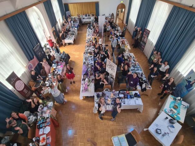 Im Inneren eines alten Saals stehen eine Reihe von Buchständen und viele Menschen, die nach oben in die Kamera schauen und winken