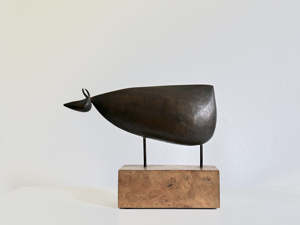 Stilisierte Plastik einer Kuh mit großem, etwas unförmigem Körper, kleinem Kopf und kleinen Stelzenbeinen auf einem Holzblock