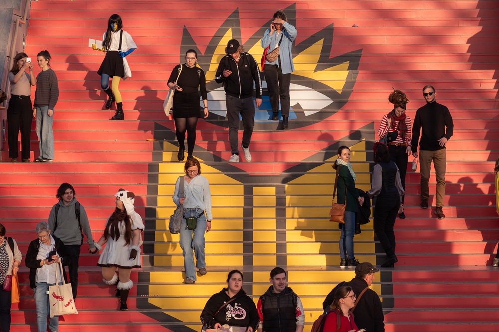 Besucher*innen der Leipziger Buchmesse gehen die Treppe hinunter, die mit dem Logo der Messe 2025 geschmückt ist: komplett rot, ein blaues Auge mit gelben Augenbrauen über gelbem Buch
