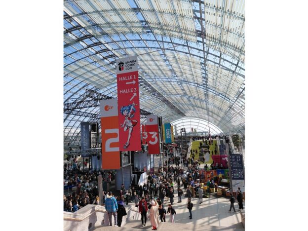 Foto vom Getümmel der Menschen auf der Leipziger Buchmesse 2026. Zu sehen ist ein großer roter Banner, der zur Manga Comic Con weißt und mehrere Banner, die Werbung für z. B. ZDF, 3 Sat oder Kika machen. Viele Menschen tummeln sich in dem gläsernen Gewölbe.