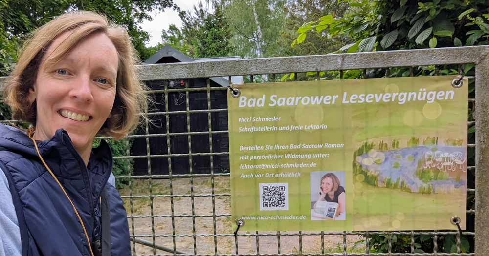 Nicci Schmieder steht vor einem Stahltor, an dem ein grünes Plakat aufgepannt ist. Auf ihm steht "Bad Saarower Lesevergnügen", darunter ein Foto von ihr, ein QR-Code und Infos zu ihr, den Büchern und Lesungen.