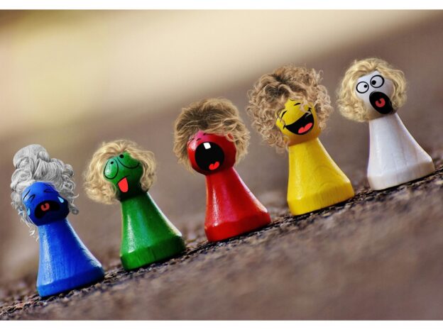 5 Holz-Spielfiguren stehen nebeneinander, alle in einer anderen Farbe, alle mit einem anderen Emoticon-Gesichtsausdruck und kleinen Perücken aufgesetzt. Links weint die blaue Figur, daneben streckt grün erschöpft die Zunge raus, rot gähnt herzhaft, gelb lacht fröhlich und weiß staunt mit offenem Mund.