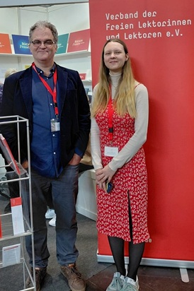 Thomas Schnakenberg und Annalena Rauh vor dem zwei Meter großen, tomatenrot leuchtenden Banner des VFLL am Stand des VFLL und der Bücherfrauen auf der Leipziger Buchmesse 2026