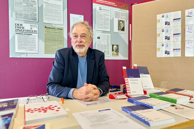 Der Verleger Klaus-Wilhelm Bramann sitzt vor einem Tisch, auf dem Infomaterial über den Verlag ausliegt sowie der Leitfaden Freies Lektorat. Er trägt einen dunkelblauen Blazer über einem hellblauen Pullover und hat die Hände auf dem Tisch gefaltet. Im Hintergrund sind auf einer leuctend pinken Wand alte Zeitungsartikel zu sehen, neben ihm steht ein Aufsteller mit Infomaterial.