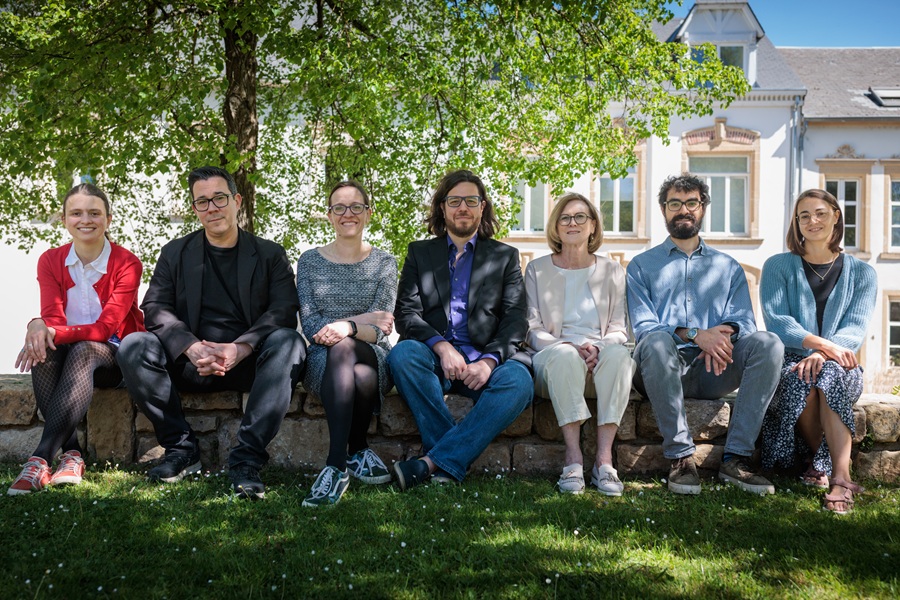 Gruppenfoto von 7 Menschen auf einem Mäuerchen vor einem grünen Baum und einem schlossähnlichen Gebäude