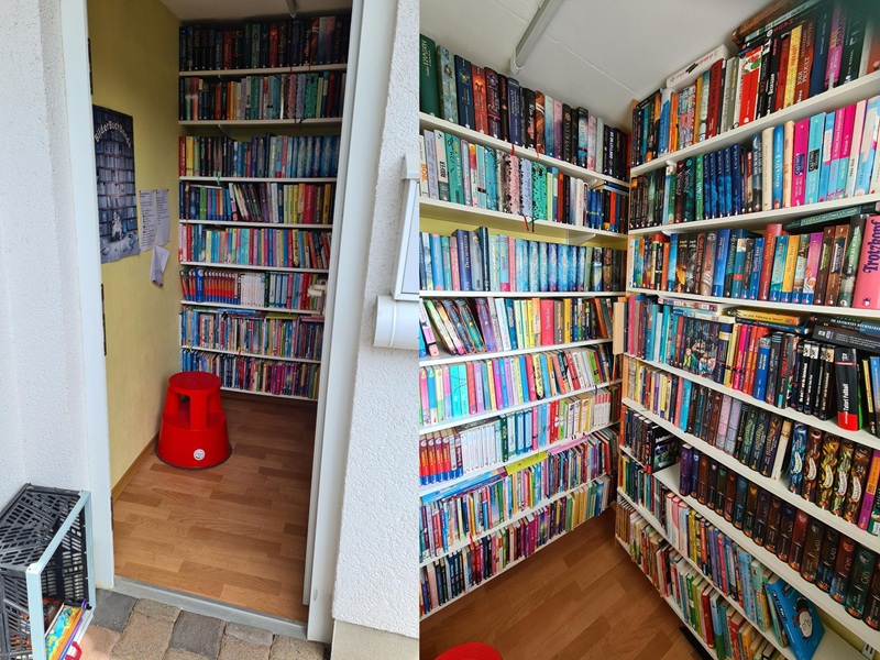 Links sieht man die Seitentür zur Garage, in der Bücherregale stehen und ein roter Plastikhocker. Unten links im Bild ist eine hochkant gestellte Kiste mit einigen Büchern daran, wohl zur Rückgabe. Im Rechten Bildsieht man, wie ca. 1 Quadratmeter der Garage abgetrennt und mit Regalen ausgestattet wurde. Vom Boden bis zur Decke sind sie prall mit Büchern gefüllt. 