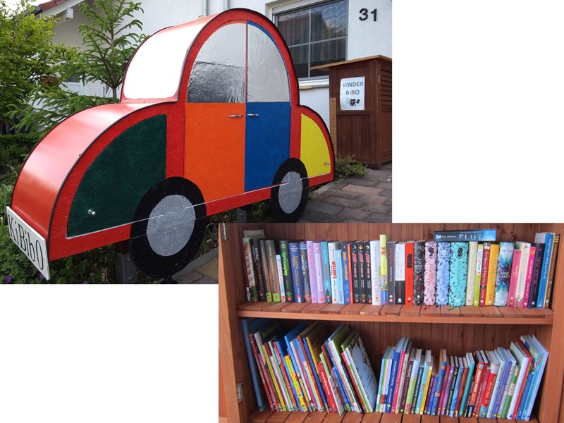 Oben das geschlossene bunte Bücherauto, ein buntes Regal in der Form eines Autos. Auf dem Nummernschild steht "Kinderbibo". Im Hintergrund sieht man einen zweiten braunen Bücherschrank mit dem Schild "Kinderbibo". Dahinter das Grün des Vorgartens.