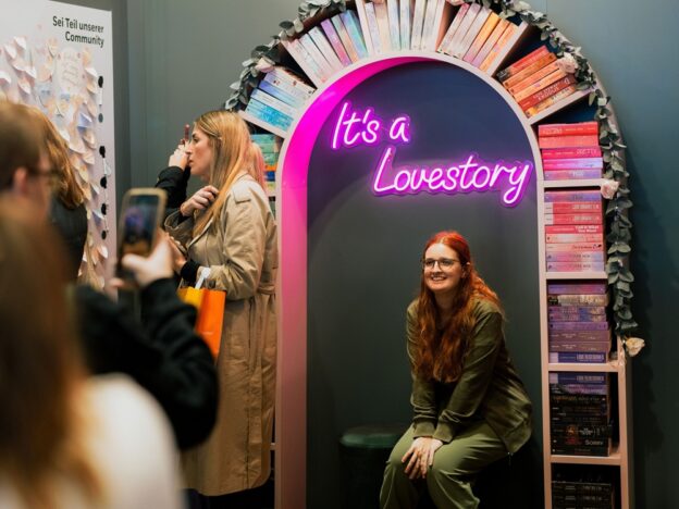 Junge Frau mit rotem Haar sitzt auf einem Hocker und schaut in einen Spiegel, den in einem Bogen bunte Bücher umgeben. Über Ihrem Gesicht im Spigel sieht man in Neonschrift: "It's a lovestory".